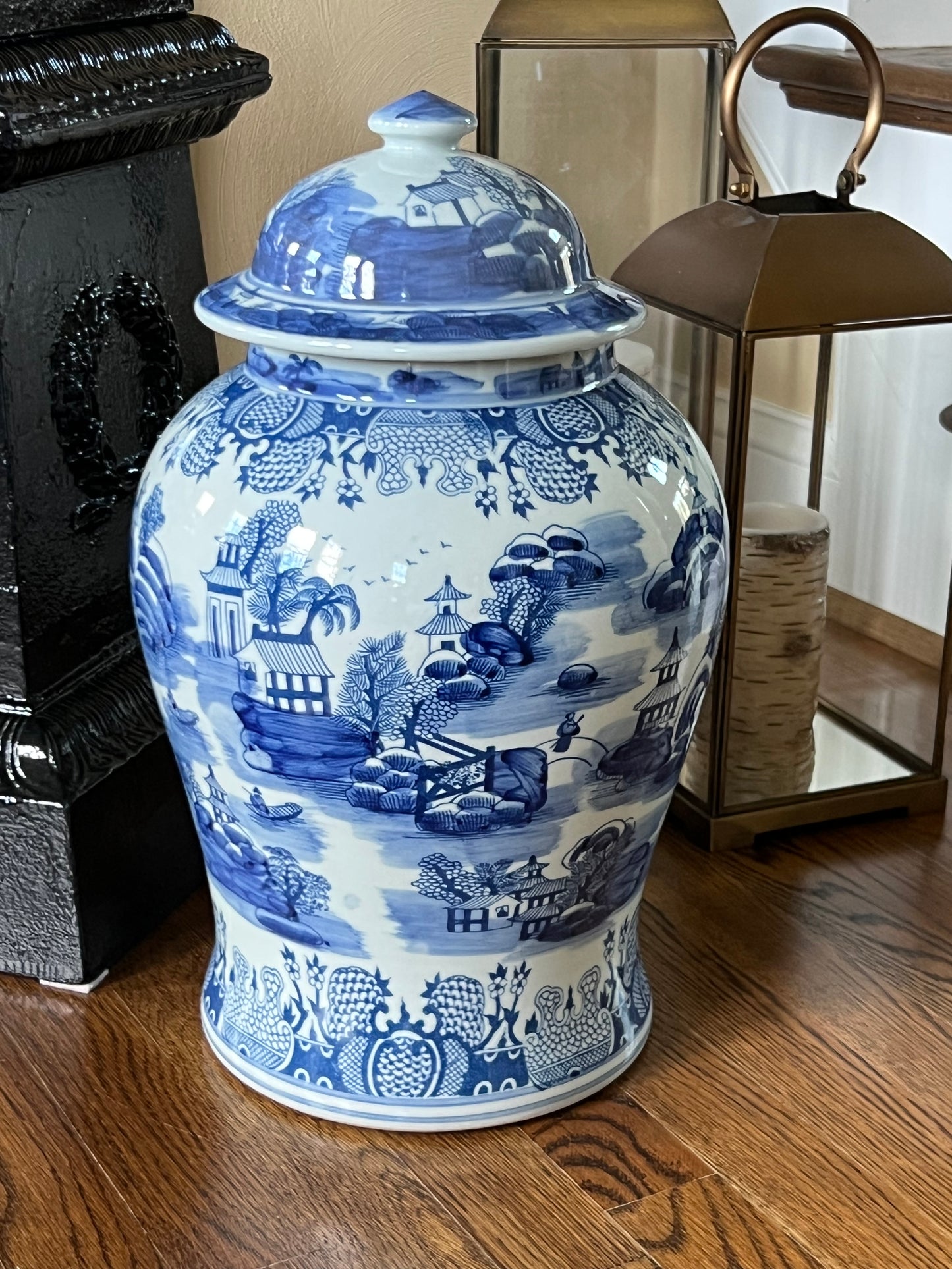 Large blue and white porcelain temple jar with pagoda landscape motif and lid, displayed on a hardwood floor in a traditional interior.