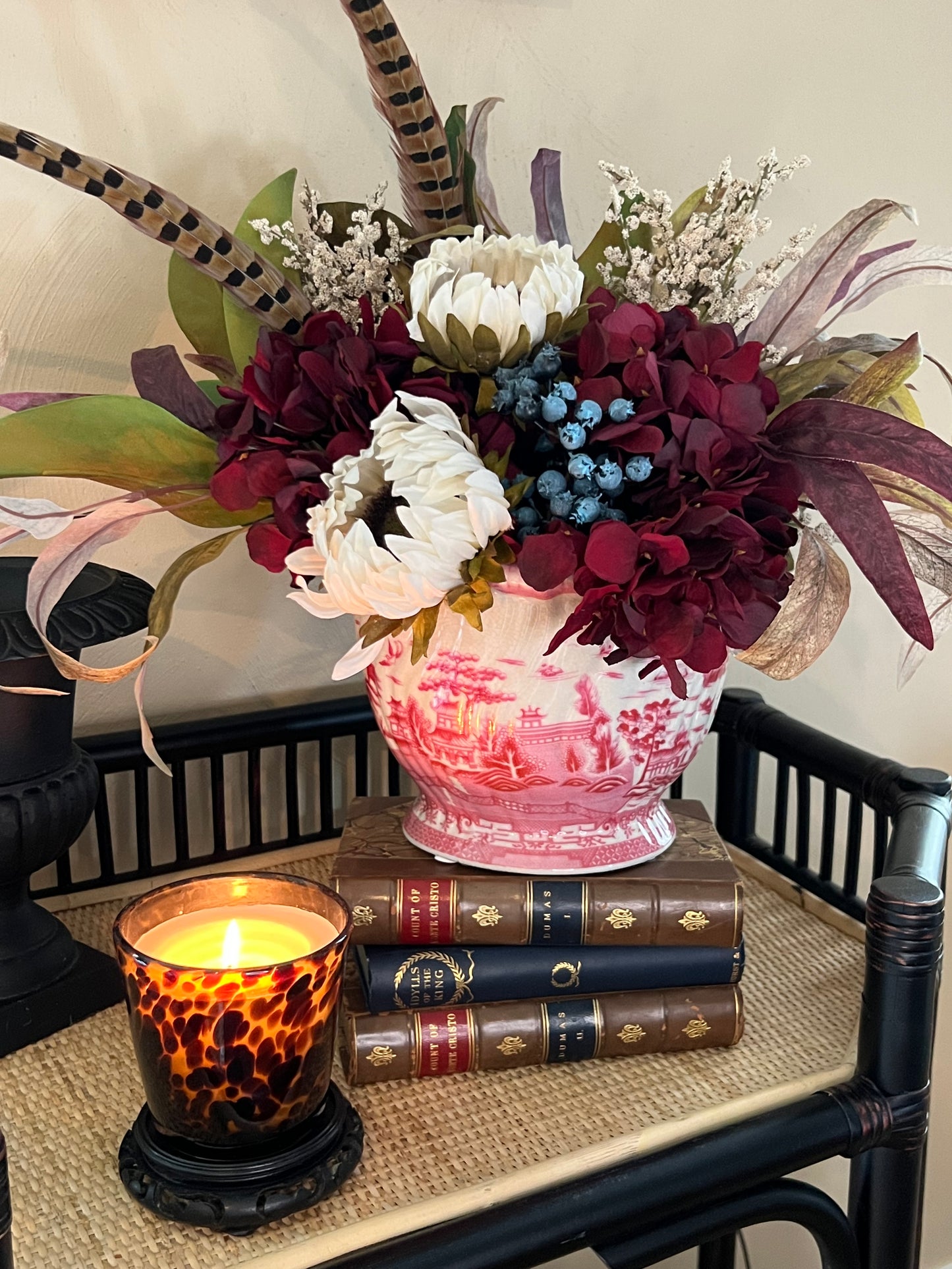 Scalloped pink pagoda chinoiserie planter filled with faux florals and pheasant feathers, styled on vintage books beside a lit tortoiseshell candle.