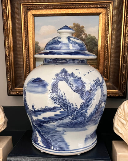 Blue and white chinoiserie landscape porcelain jar with lid on a dark surface with framed artwork in the background