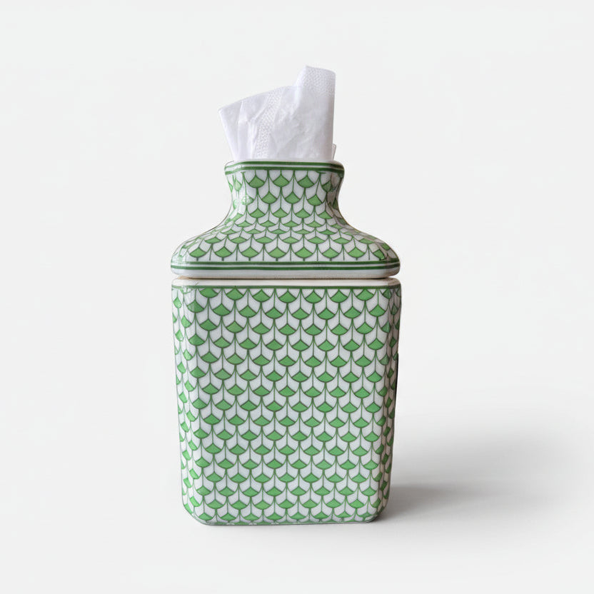 Green and white fish scale porcelain tissue box cover with tissues inserted, styled on a dark tabletop with a gold framed artwork and white flowers in the background.
