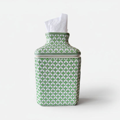 Green and white fish scale porcelain tissue box cover with tissues inserted, styled on a dark tabletop with a gold framed artwork and white flowers in the background.