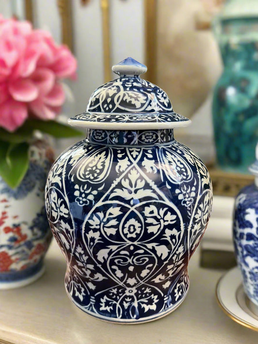 Cobalt blue and white porcelain temple jar with damask pattern and matching lid, styled on a tabletop with pink florals in the background.