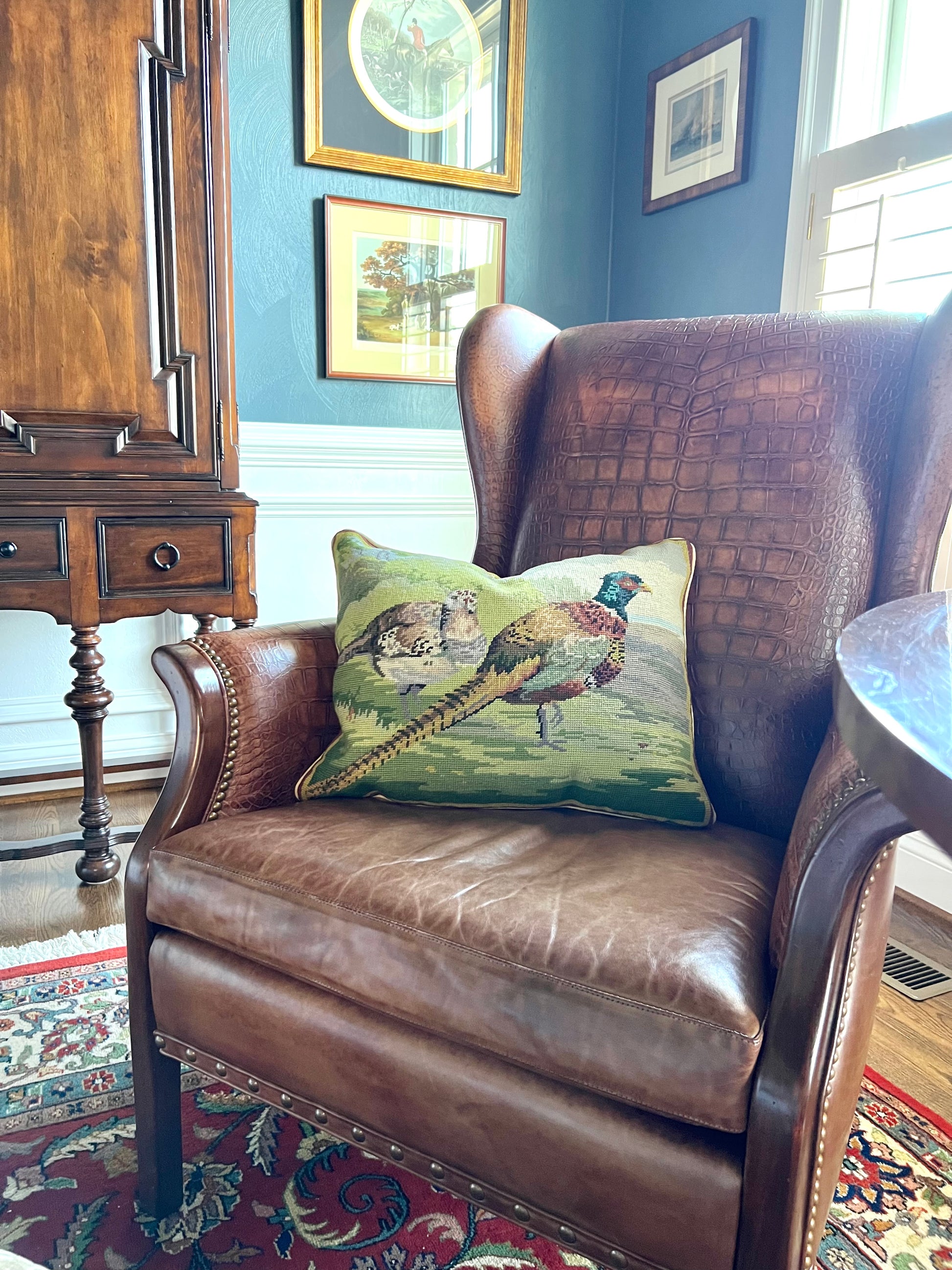 Needlepoint pheasant and grouse pillow styled on a brown leather wingback chair in a traditional room.