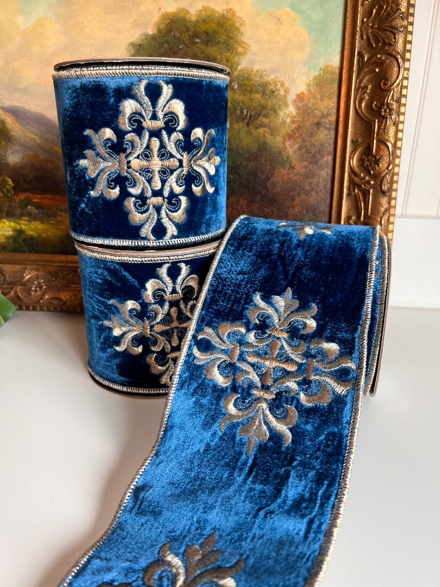 A roll of royal blue velvet ribbon with gold embroidered medallion motifs, displayed next to a matching roll on a white surface with a framed painting in the background.