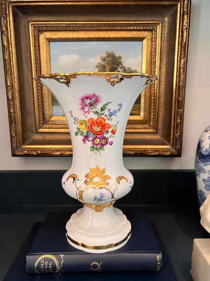Meissen B-Form gold encrusted hand painted floral porcelain krater vase styled on top of a dark book against a gold frame.