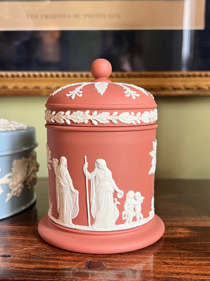 A classic Wedgwood-style ceramic lidded jar in terracotta with white relief figures and decorative borders, displayed on a wooden table.