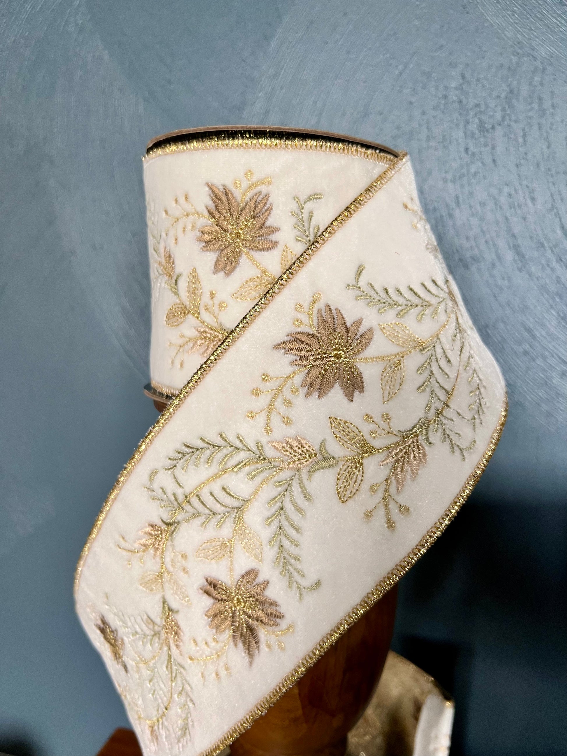 A roll of winter white velvet ribbon with gold and soft bronze floral embroidery and a thin gold metallic edge, displayed on a wooden stand against a textured blue-gray background.