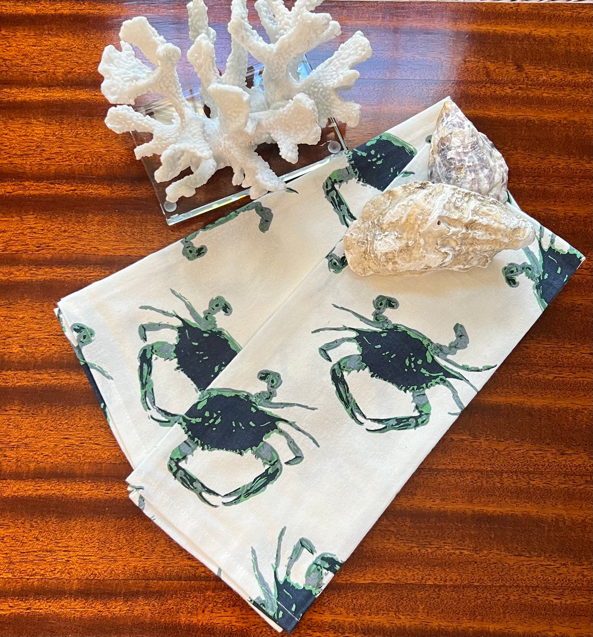 Green crab print tea towels styled with coral and an oyster shell on a wood surface.