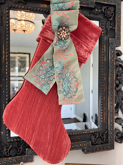 Coral crushed velvet Christmas stocking styled with vintage brooch and decorative ribbon hanging from an ornate decorative mirror.