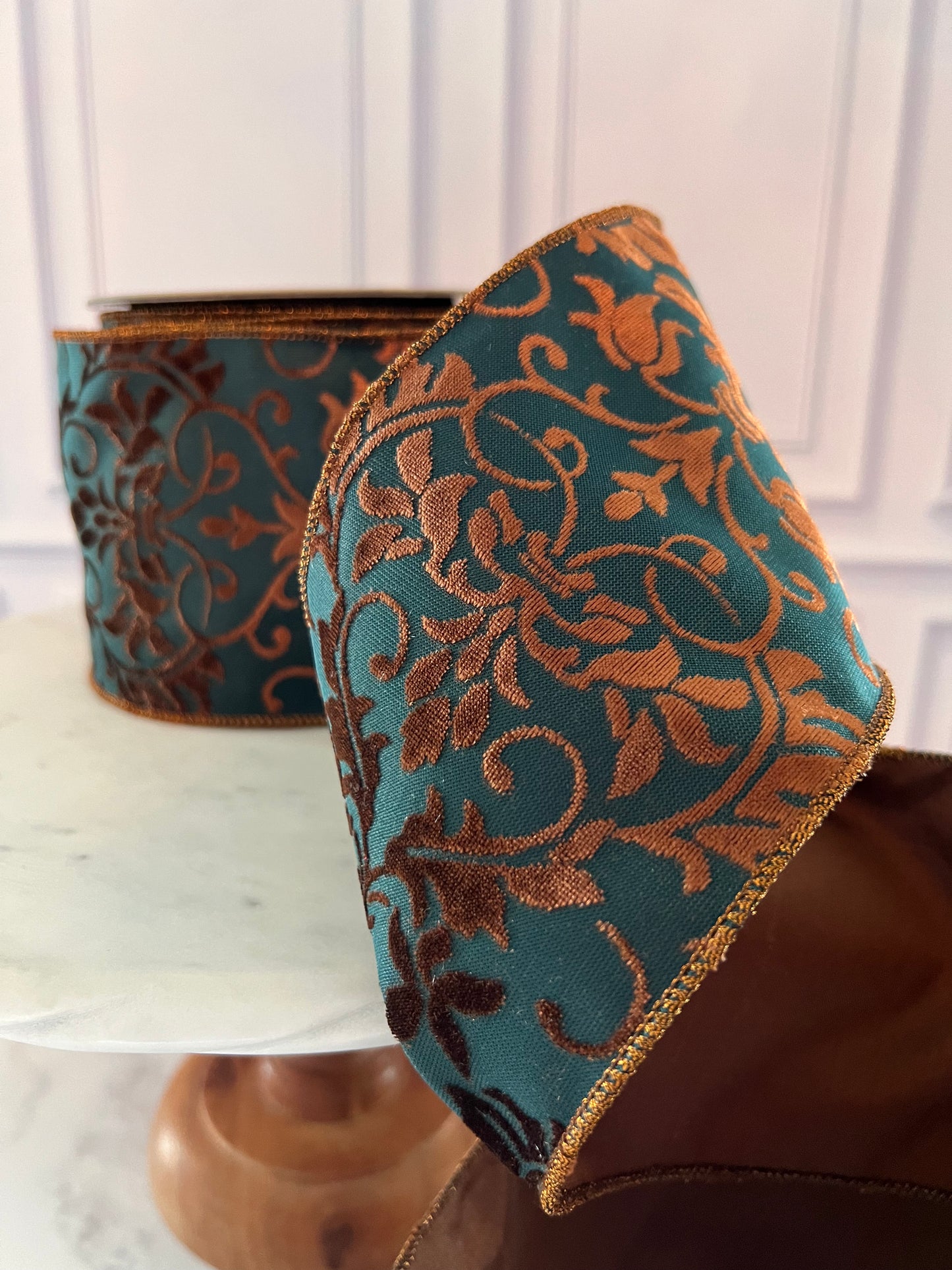 A roll of teal and copper-brown damask ribbon with gold trim, shown in a close-up with the unrolled section draped over a brown surface.