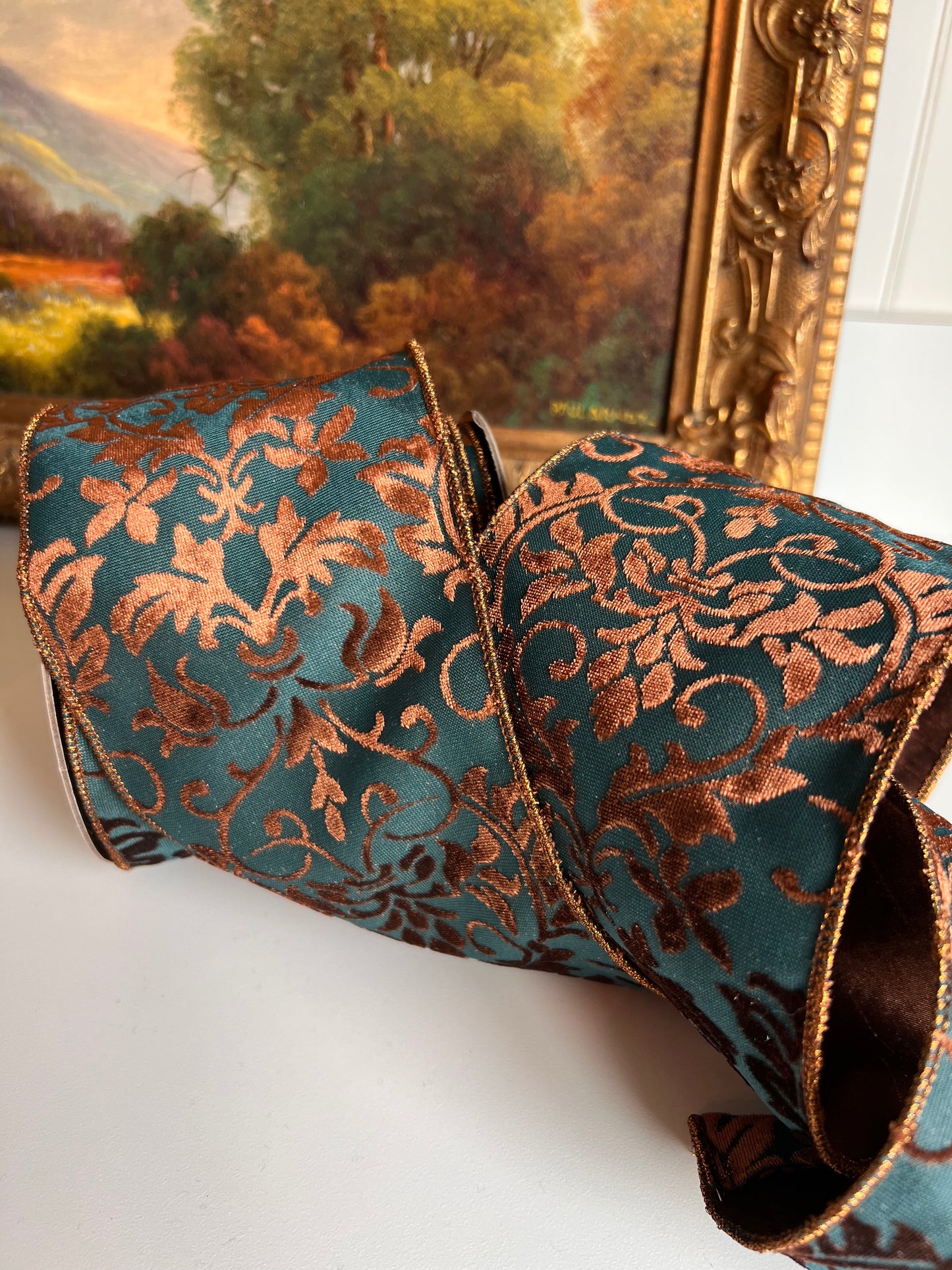 A roll of decorative ribbon with a teal blue background and metallic copper-brown damask floral pattern, displayed on a white surface with a framed landscape painting in the background.