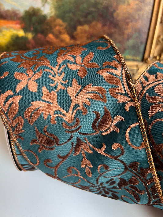 A close-up of a roll of decorative ribbon with a teal blue background and a metallic copper-brown damask floral pattern, finished with a thin metallic edge trim.
